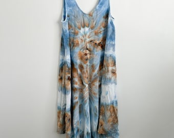 May include: A sleeveless midi dress featuring a tie-dye pattern in shades of light blue, white, and earthy brown. The garment has a V-neckline, a relaxed fit, and an asymmetrical hemline that falls lower at the sides, hanging on a wire hanger.
