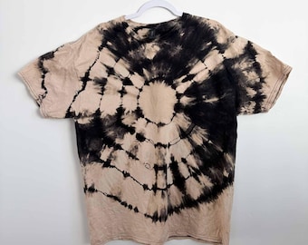 May include: A short-sleeve crew neck t-shirt featuring a tie-dye pattern with a circular, sunburst-like design in black and beige. The black dye radiates from the center in distinct, segmented lines against the light beige fabric background.