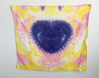 May include: A square fabric bandana featuring a tie-dye pattern with a large, dark purple heart shape centered on the material. The surrounding area displays a vibrant blend of bright yellow and soft pink tie-dye swirls across the entire surface.