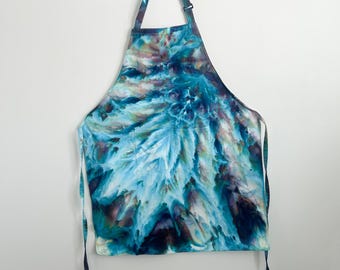 May include: A tie-dye apron featuring a vibrant color palette of deep navy, bright turquoise, and soft lavender in a swirling, abstract pattern. The apron includes an adjustable neck strap with a plastic slider and two long side ties.