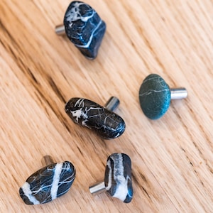 Può includere: Una collezione di sei pomelli per mobili in pietra con steli color argento. I pomelli sono di varie forme e dimensioni, con motivi di venature nere, bianche e grigie. I pomelli sono esposti su una superficie di legno chiaro.