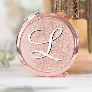 May include: A rose gold compact mirror with a sparkly pink glitter surface and a white cursive letter "L" in the centre.