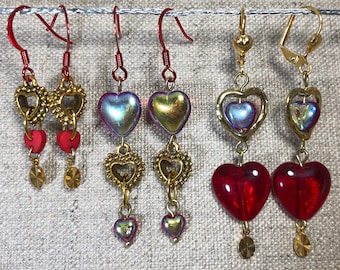 May include: Three pairs of gold heart earrings with red glass beads. The earrings are shown in three lengths: short, medium, and long. The earrings are all made with gold-toned metal and have a heart-shaped design.