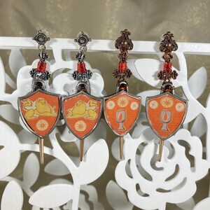 May include: Three pairs of dangle earrings with a sword design. Each earring features a shield with a different design: two lions, a goblet, and a goblet. The shields are orange with gold accents. The swords are silver with red beads.