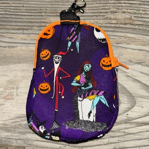 Puede incluir: Una peque&ntilde;a bolsa morada con cremallera con personajes y calabazas naranjas de The Nightmare Before Christmas. La bolsa tiene una cremallera y ribete naranja, una correa negra y un cierre plateado.