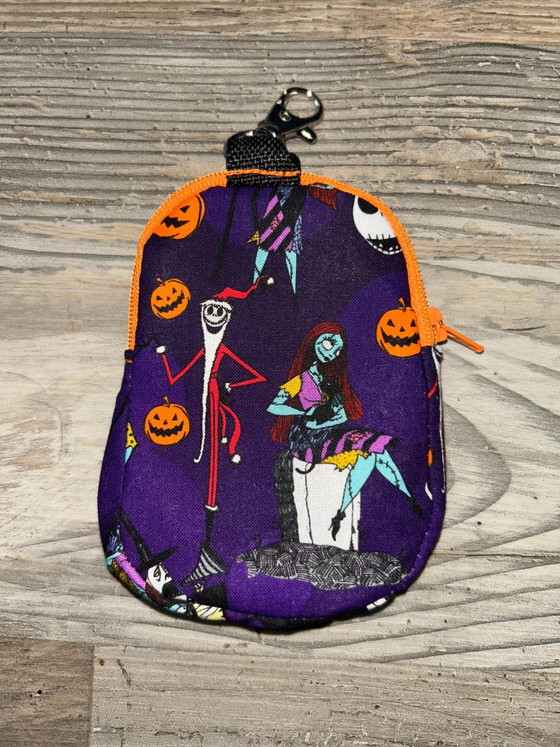 Puede incluir: Una peque&ntilde;a bolsa morada con cremallera con personajes y calabazas naranjas de The Nightmare Before Christmas. La bolsa tiene una cremallera y ribete naranja, una correa negra y un cierre plateado.