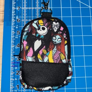 May include: A small backpack-shaped pouch featuring a print of Jack Skellington and Sally. The pouch has a black mesh front pocket, a black zipper, and a metal lobster clasp at the top. It measures approximately 5.5 inches in height and 3.5 inches in width.