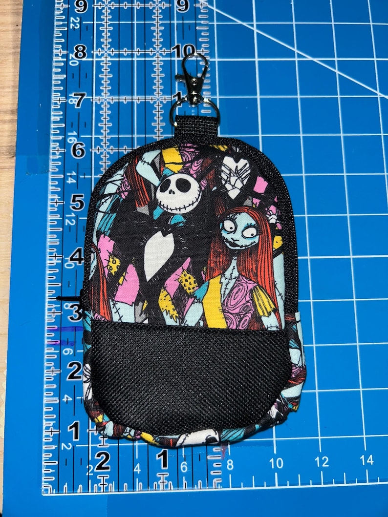 May include: A small backpack-shaped pouch featuring a print of Jack Skellington and Sally. The pouch has a black mesh front pocket, a black zipper, and a metal lobster clasp at the top. It measures approximately 5.5 inches in height and 3.5 inches in width.