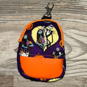 Puede incluir: Peque&ntilde;o estuche morado con cremallera, con un acento naranja y un cierre plateado. El estuche presenta una impresi&oacute;n de personajes de Pesadilla antes de Navidad, incluidos Jack Skellington y Sally. Los bordes superior e inferior son de color naranja.
