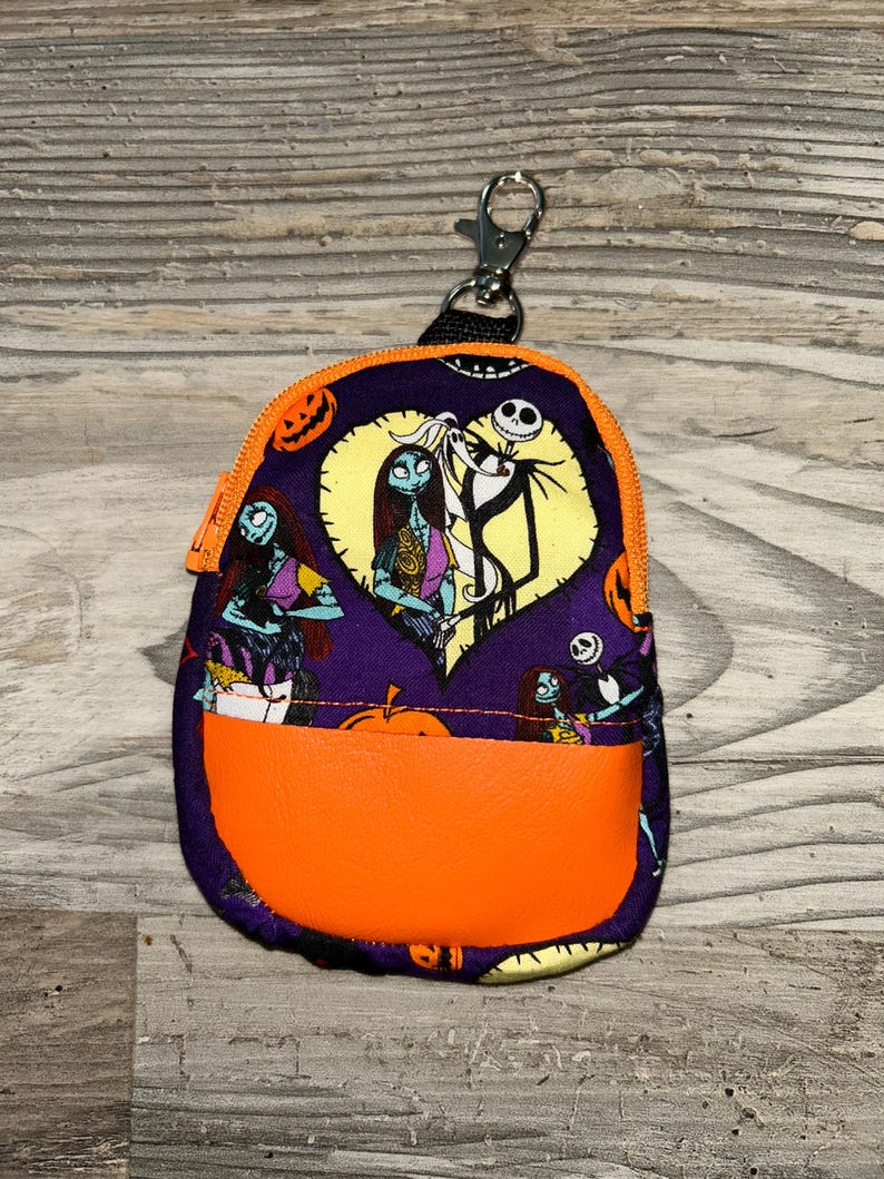 Puede incluir: Peque&ntilde;o estuche morado con cremallera, con un acento naranja y un cierre plateado. El estuche presenta una impresi&oacute;n de personajes de Pesadilla antes de Navidad, incluidos Jack Skellington y Sally. Los bordes superior e inferior son de color naranja.