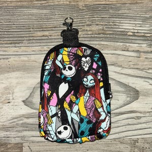 May include: A small, rectangular zippered pouch featuring a colorful, repeating pattern of Jack Skellington and Sally from The Nightmare Before Christmas. The pouch includes a black fabric border, a black zipper, and a metal lobster claw clasp.