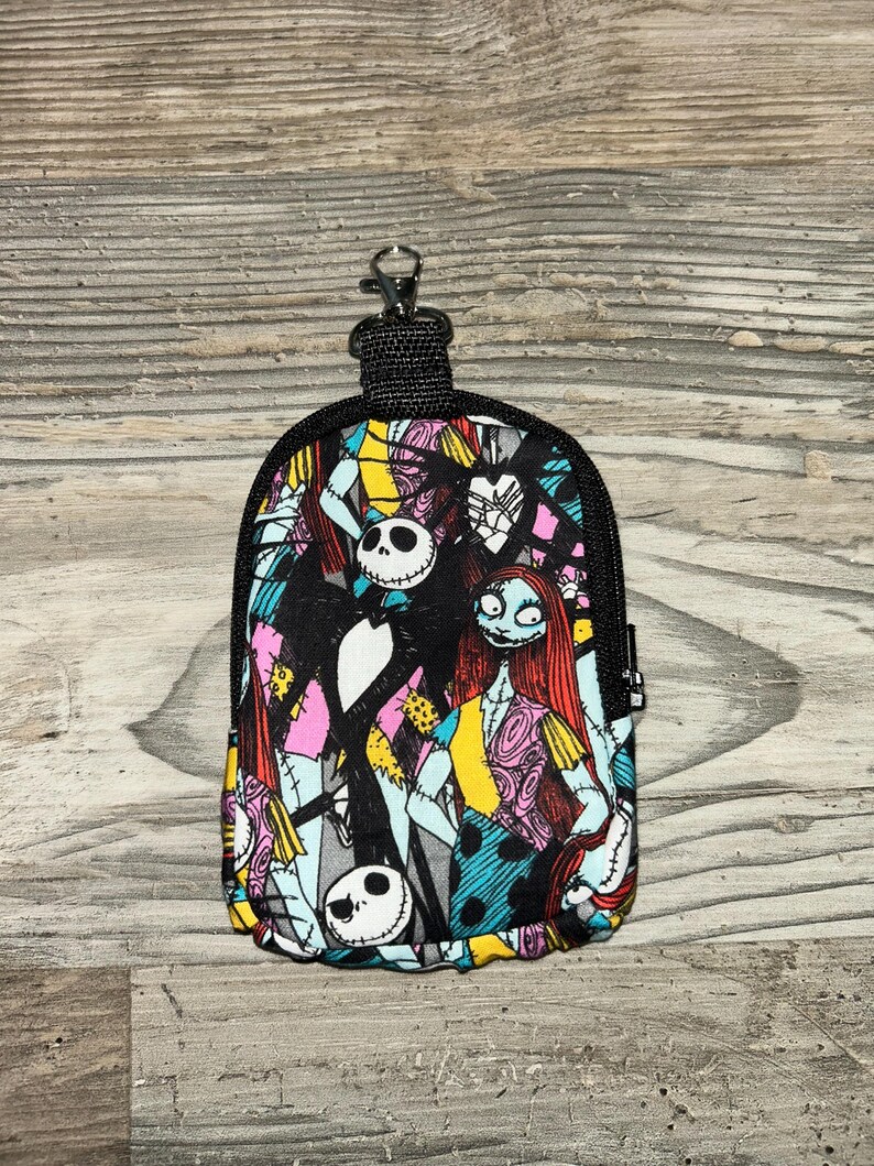 May include: A small, rectangular zippered pouch featuring a colorful, repeating pattern of Jack Skellington and Sally from The Nightmare Before Christmas. The pouch includes a black fabric border, a black zipper, and a metal lobster claw clasp.