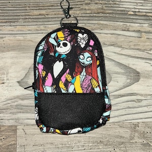 May include: A small backpack-shaped pouch featuring a colorful print of Jack Skellington and Sally from The Nightmare Before Christmas. The pouch has a black mesh front pocket, a black zipper closure, and a silver metal lobster clasp at the top.
