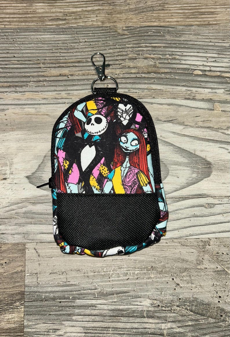 May include: A small backpack-shaped pouch featuring a colorful print of Jack Skellington and Sally from The Nightmare Before Christmas. The pouch has a black mesh front pocket, a black zipper closure, and a silver metal lobster clasp at the top.