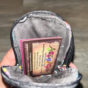 May include: A small, round, zippered pouch with a dark gray interior lining. Inside the pouch is a rectangular business card with the text Artistic Attitude, featuring a wood-grain background and colorful paint splatter accents.