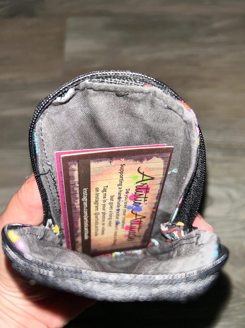 May include: A small, round, zippered pouch with a dark gray interior lining. Inside the pouch is a rectangular business card with the text Artistic Attitude, featuring a wood-grain background and colorful paint splatter accents.