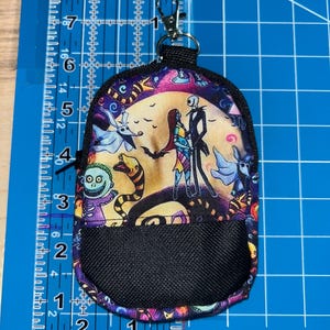 May include: A small fabric pouch featuring a colorful print of characters from The Nightmare Before Christmas, including Jack Skellington and Sally. The pouch has a black mesh front pocket, a black zipper, and a silver metal lobster claw clasp.