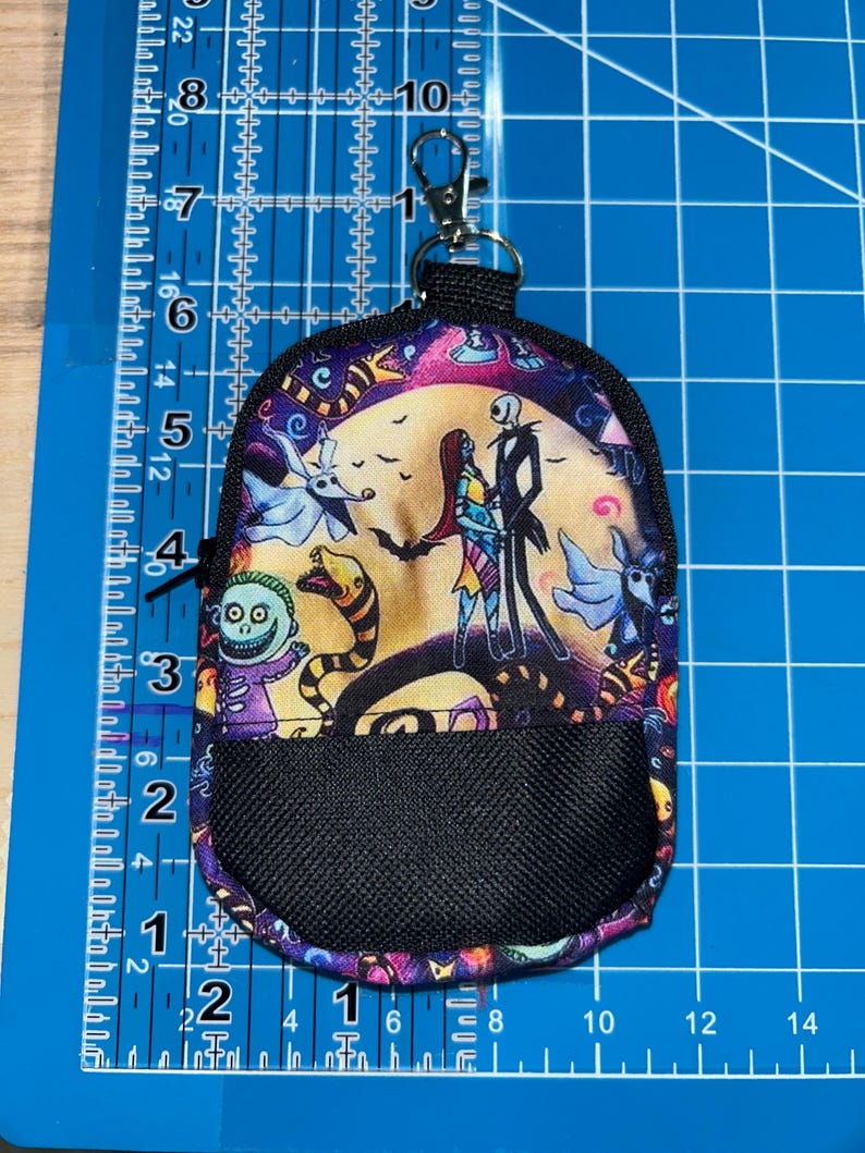 May include: A small fabric pouch featuring a colorful print of characters from The Nightmare Before Christmas, including Jack Skellington and Sally. The pouch has a black mesh front pocket, a black zipper, and a silver metal lobster claw clasp.