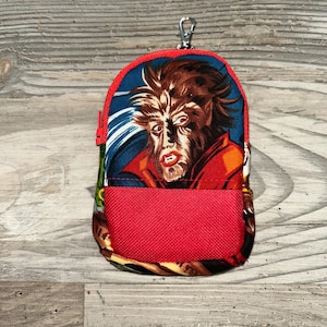 Może przedstawiać: Mała, czerwono-niebieska saszetka zapinana na zamek z grafiką wilkołaka. Saszetka posiada czerwoną kieszeń z siatki i srebrny klips. Grafika wilkołaka znajduje się na środku saszetki.