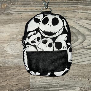 May include: A small, rectangular zippered pouch featuring a repeating black and white pattern of Jack Skellington faces. The pouch includes a black mesh front pocket and a silver-toned metal lobster clasp attached to the top for easy hanging.