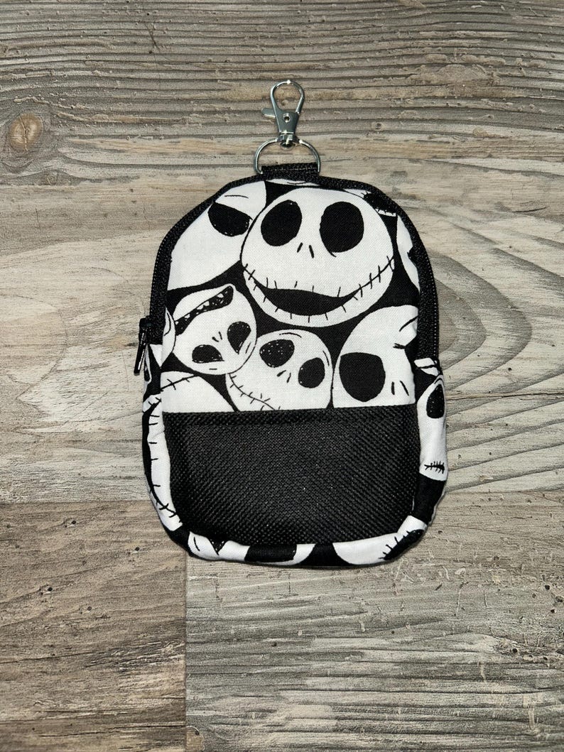 May include: A small, rectangular zippered pouch featuring a repeating black and white pattern of Jack Skellington faces. The pouch includes a black mesh front pocket and a silver-toned metal lobster clasp attached to the top for easy hanging.