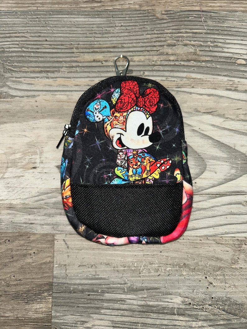 May include: A small black zipped pouch featuring a colourful graphic of Minnie Mouse wearing a red bow, surrounded by various Disney character illustrations and a star-patterned background. The pouch includes a black mesh front pocket and a metal clip.