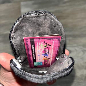 May include: A small, dark gray, textured fabric pouch with a black zipper closure. Inside the pouch is a rectangular business card with a pink and black background, featuring the text Artistic Attitude and steffaniesews.etsy.com.