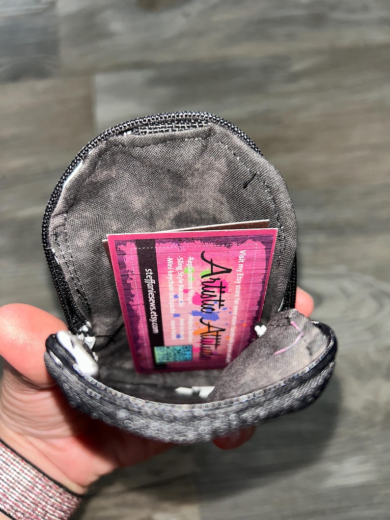 May include: A small, dark gray, textured fabric pouch with a black zipper closure. Inside the pouch is a rectangular business card with a pink and black background, featuring the text Artistic Attitude and steffaniesews.etsy.com.