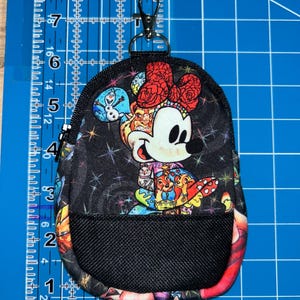 May include: A small black fabric pouch with a zip closure and a metal lobster clasp. The pouch features a colourful print of cartoon characters, including a mouse with a red bow, against a starry background, with a black mesh pocket on the front.