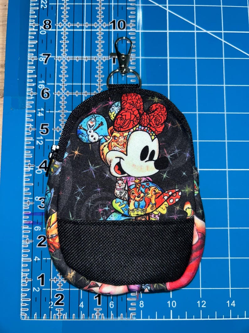 May include: A small black fabric pouch with a zip closure and a metal lobster clasp. The pouch features a colourful print of cartoon characters, including a mouse with a red bow, against a starry background, with a black mesh pocket on the front.