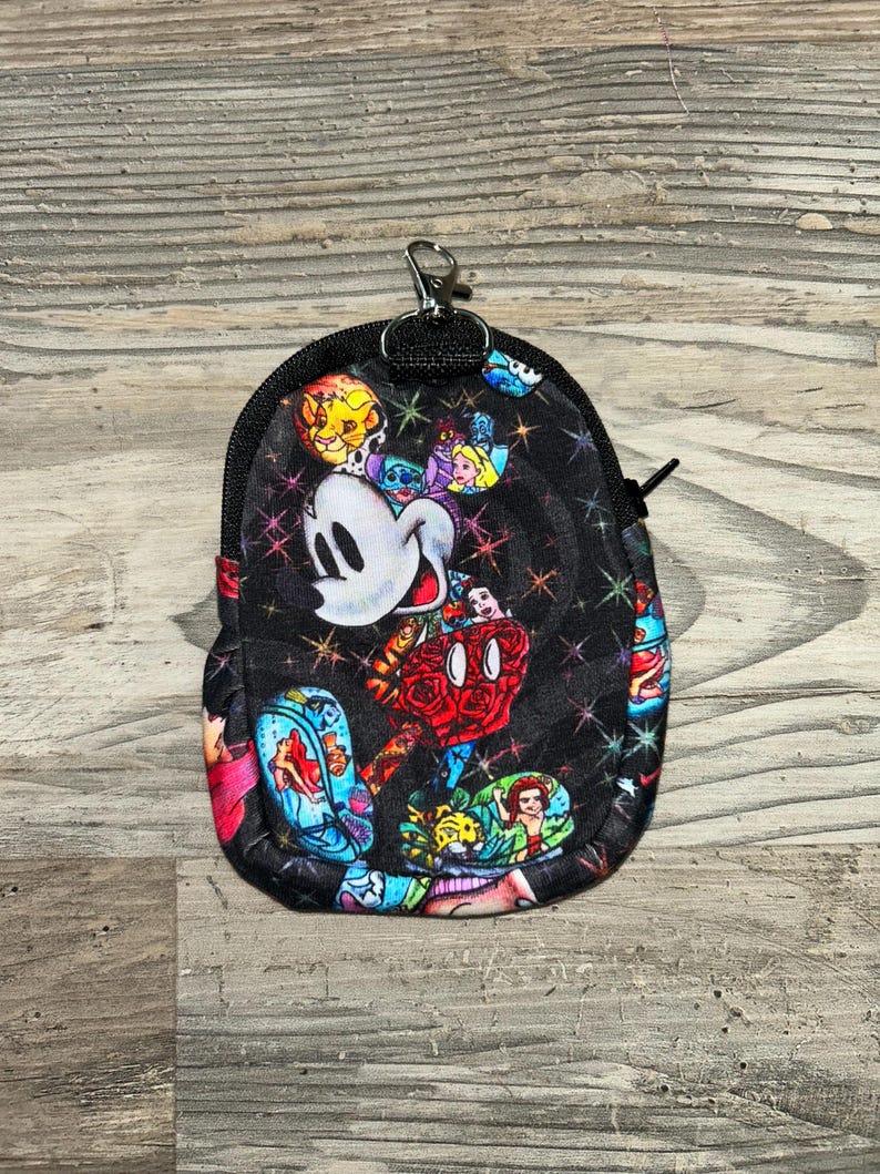 May include: A small black zippered pouch featuring a colourful all-over print of various animated characters, including a prominent white mouse head, surrounded by small multicoloured starbursts. The pouch includes a silver metal lobster clasp.