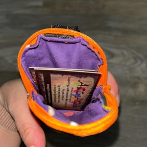Puede incluir: Una peque&ntilde;a bolsa naranja con cremallera y un interior morado. La bolsa contiene una peque&ntilde;a tarjeta con el texto "Artistic Attic". La bolsa est&aacute; abierta, revelando el forro interior y la tarjeta. La cremallera es negra.