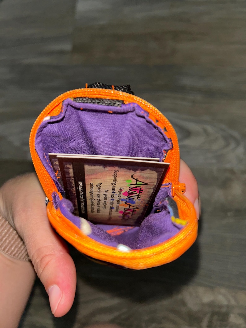 Puede incluir: Una peque&ntilde;a bolsa naranja con cremallera y un interior morado. La bolsa contiene una peque&ntilde;a tarjeta con el texto "Artistic Attic". La bolsa est&aacute; abierta, revelando el forro interior y la tarjeta. La cremallera es negra.