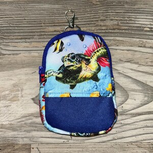 May include: Miniature backpack keychain with a blue, sea-themed design. The fabric features a sea turtle, fish, and coral against a blue background. The backpack has a blue mesh pocket and a silver-colored clasp.
