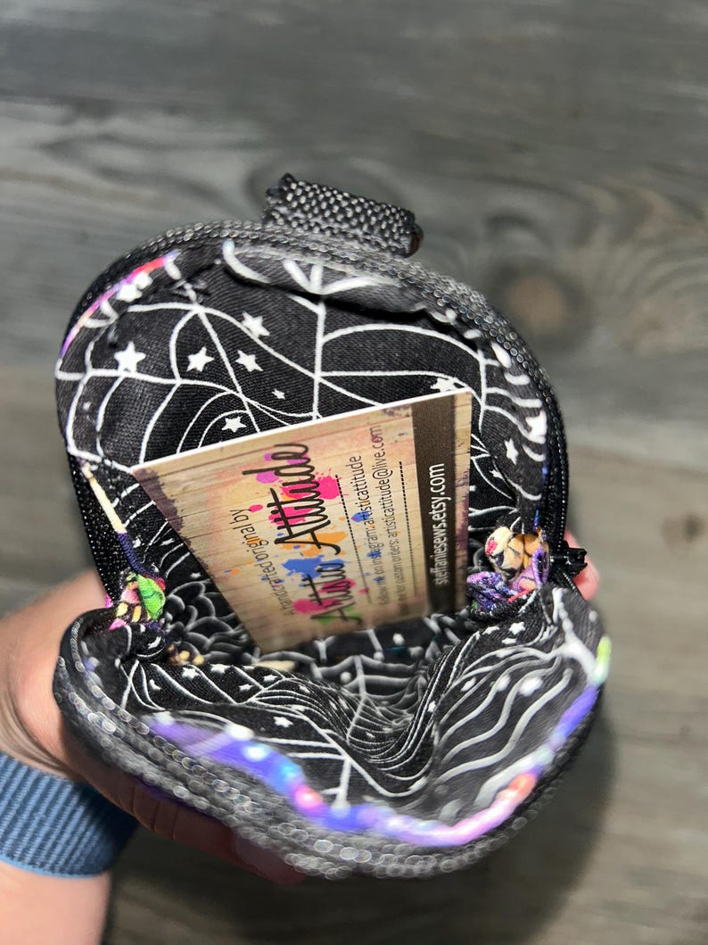 May include: A small, zippered pouch with a black interior lining featuring a white spiderweb and star pattern. Inside the pouch is a rectangular business card with the text Artistic Attitude, social media handles, and steffaniesews.etsy.com.