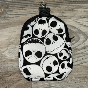 May include: A small, rounded pouch featuring a repeating black and white pattern of Jack Skellington faces. The pouch has a black zipper closure along the top edge and includes a silver-toned metal lobster clasp attached to a black fabric loop.
