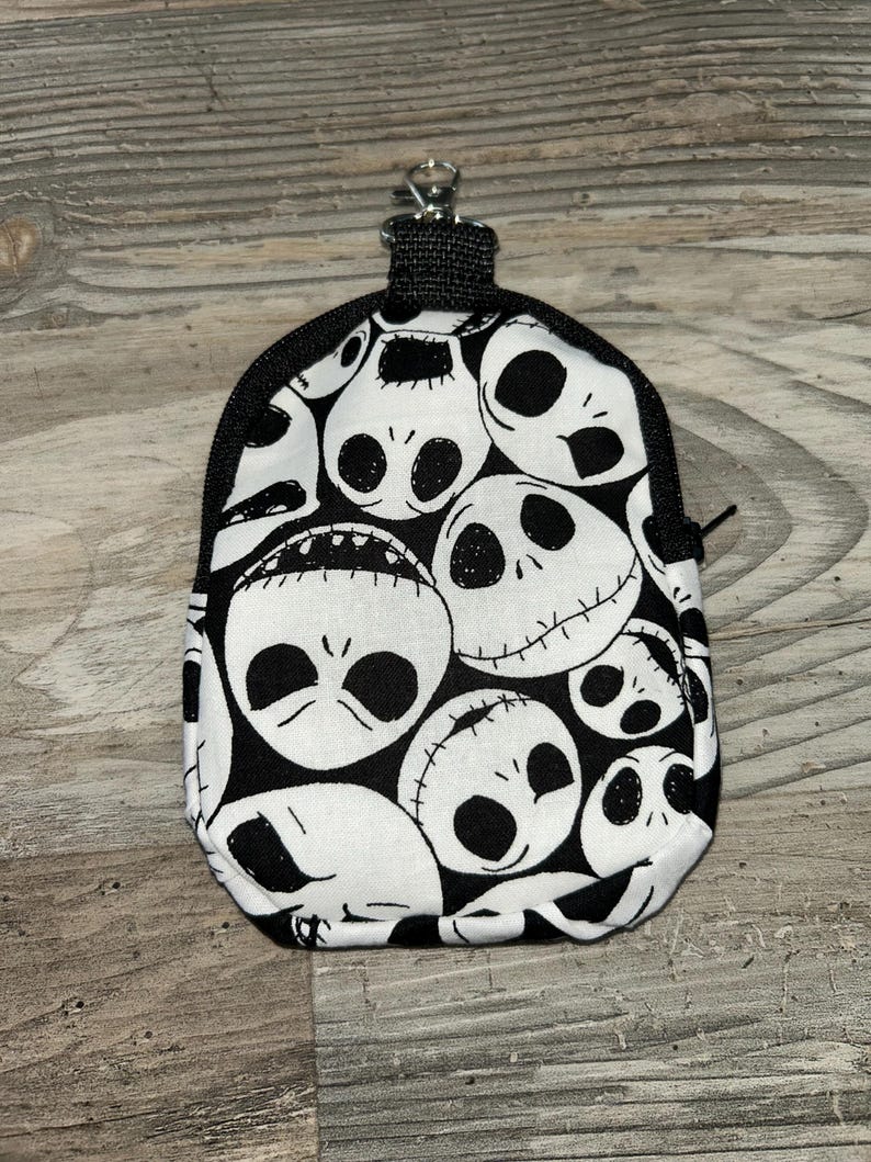 May include: A small, rounded pouch featuring a repeating black and white pattern of Jack Skellington faces. The pouch has a black zipper closure along the top edge and includes a silver-toned metal lobster clasp attached to a black fabric loop.
