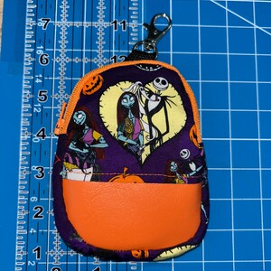 Puede incluir: Un peque&ntilde;o monedero morado con forma de mochila con cremallera y bolsillo naranja. El monedero presenta un estampado tem&aacute;tico de Halloween con Jack y Sally de Pesadilla antes de Navidad. Se adjunta un cierre plateado.