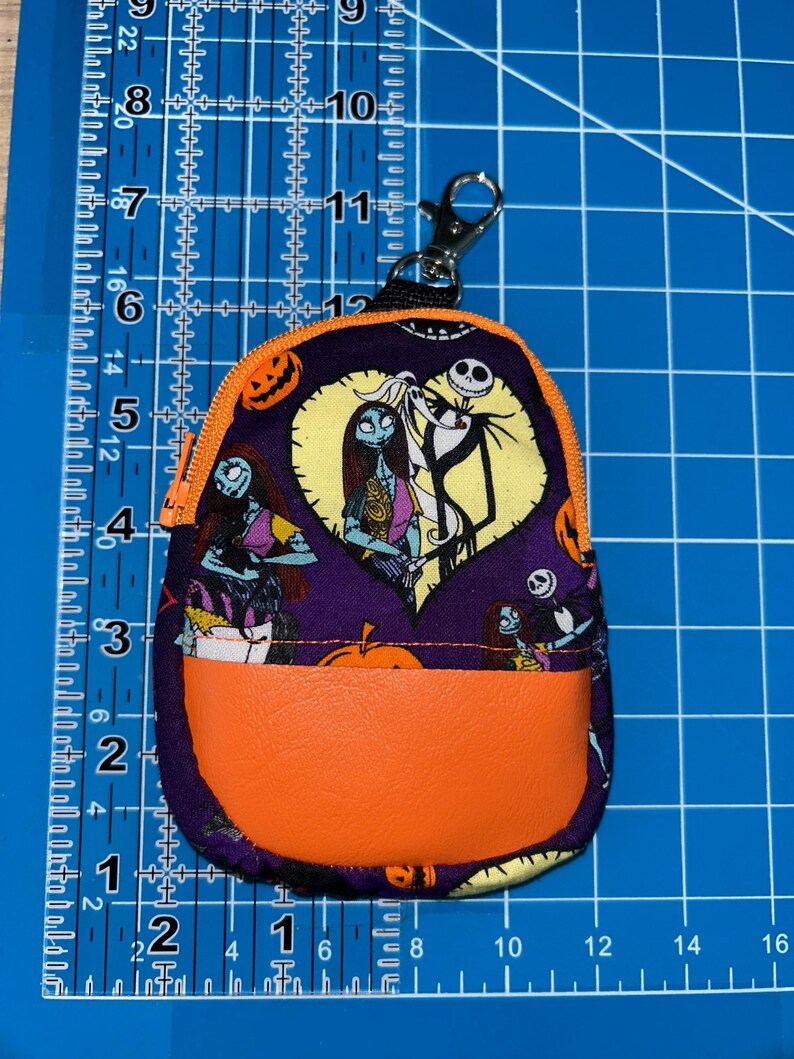 Puede incluir: Un peque&ntilde;o monedero morado con forma de mochila con cremallera y bolsillo naranja. El monedero presenta un estampado tem&aacute;tico de Halloween con Jack y Sally de Pesadilla antes de Navidad. Se adjunta un cierre plateado.