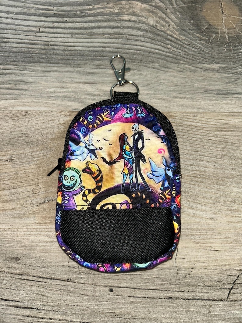 May include: A small, rounded zipper pouch featuring a colorful print of characters from The Nightmare Before Christmas, including Jack Skellington and Sally. The pouch has a black mesh front pocket, black trim, and a silver metal lobster claw clasp.