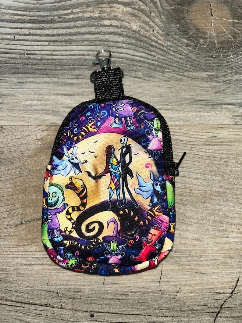 May include: A small zippered pouch featuring a colorful print of characters from The Nightmare Before Christmas, including Jack Skellington and Sally standing before a large yellow moon, with a black fabric trim and a metal lobster claw clasp.