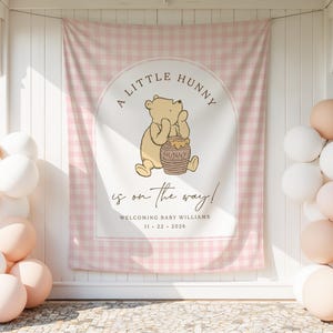 May include: A rectangular fabric backdrop featuring a light pink and white gingham pattern with a central arch containing a cartoon bear holding a honey pot. Text reads A LITTLE HUNNY is on the way! WELCOMING BABY WILLIAMS 11 22 2026.