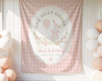 May include: A pink gingham backdrop with a central oval design featuring a goose wearing a party hat and holding a balloon. Text reads "One Silly Goose" and "Lillian's 1st Birthday, April 6th, 2025". Balloons are on the left and right.