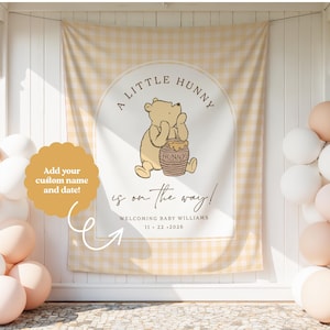 May include: A rectangular fabric backdrop featuring a light yellow gingham pattern with a central arch design. It displays a cartoon bear holding a honey pot, with the text A LITTLE HUNNY is on the way! WELCOMING BABY WILLIAMS 11 22 2026.