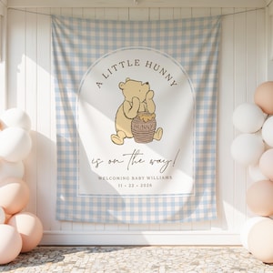 May include: A rectangular fabric backdrop featuring a light blue and white gingham pattern with a central arch design. It displays a yellow bear illustration holding a honey pot, with the text A LITTLE HUNNY is on the way! WELCOMING BABY WILLIAMS 11 22 2026.
