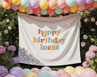 May include: A white banner with the words "happy birthday lucas" in colorful retro font is displayed outdoors. The banner is surrounded by colorful balloons and lush greenery, creating a festive atmosphere. The balloons are in shades of pink, yellow, and blue.