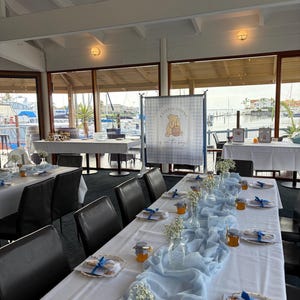 May include: A long dining table set for an event with a white tablecloth, a light blue fabric runner, small glass vases with baby's breath, and small jars of honey with blue and white checkered lids. A banner reads A LITTLE HUNNY is on the way.