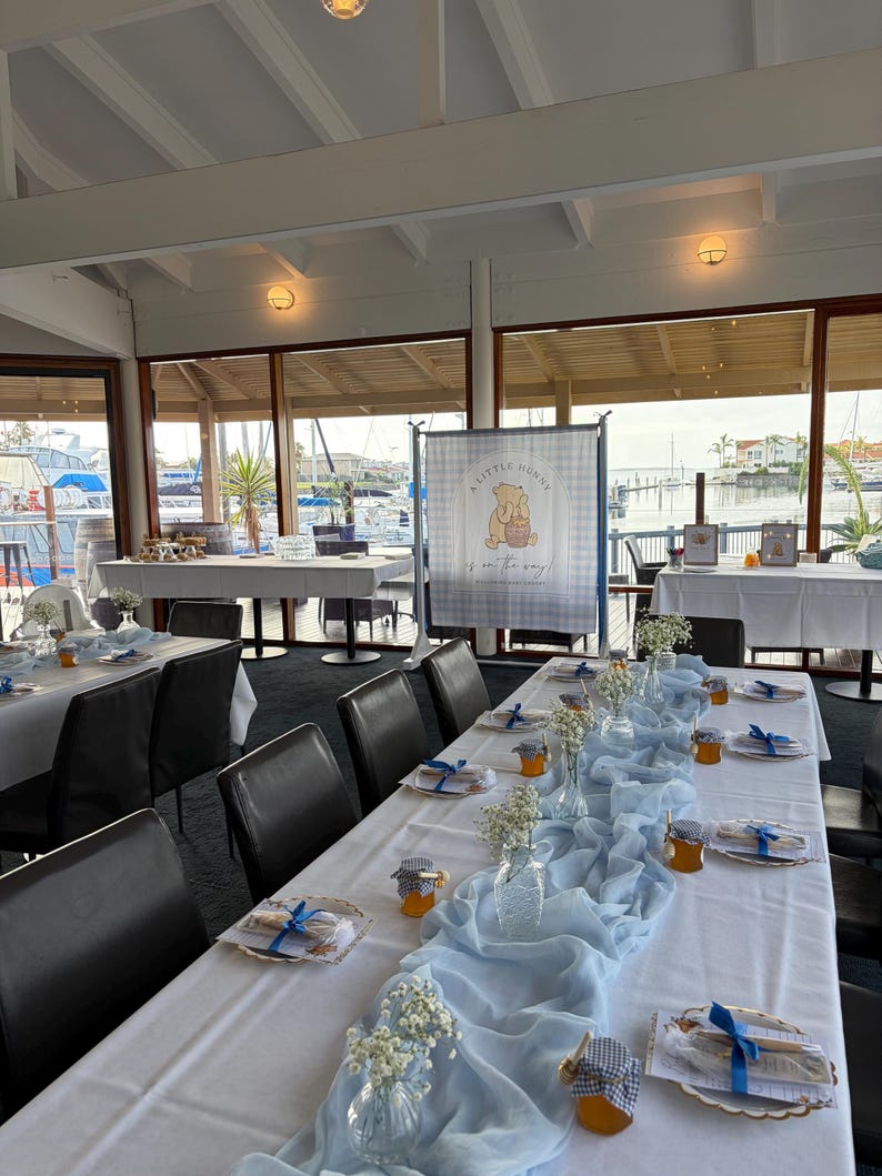 May include: A long dining table set for an event with a white tablecloth, a light blue fabric runner, small glass vases with baby's breath, and small jars of honey with blue and white checkered lids. A banner reads A LITTLE HUNNY is on the way.