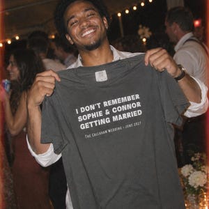 Op de afbeelding: Een donkergrijs t-shirt met korte mouwen en witte tekst: I DON'T REMEMBER SOPHIE & CONNOR GETTING MARRIED, gevolgd door de tekst: THE CALLAHAN WEDDING - JUNE 2027. Het t-shirt heeft een zichtbaar merklabel aan de binnenkant van de kraag.