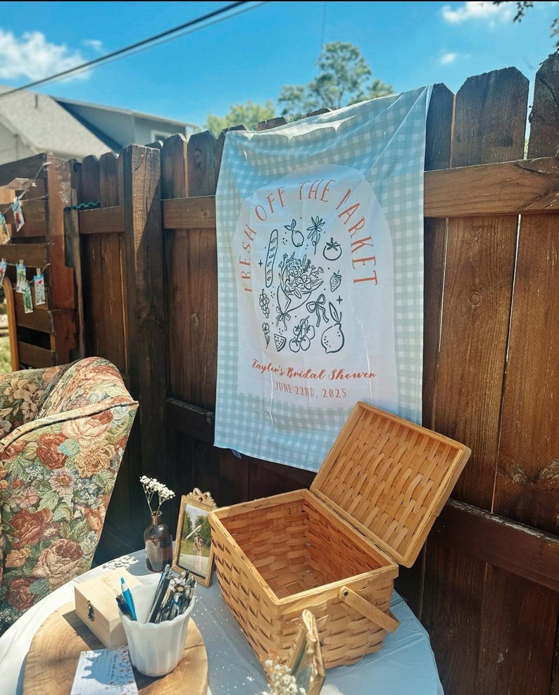 May include: A picnic scene with a woven basket, a sign that says "Fresh Off The Market", and a floral patterned chair. The sign is hanging on a wooden fence. The scene is set outdoors.